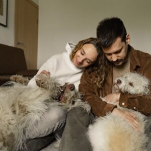 A joyful couple hugging their two fluffy dogs while sitting indoors, showcasing love and companionship.